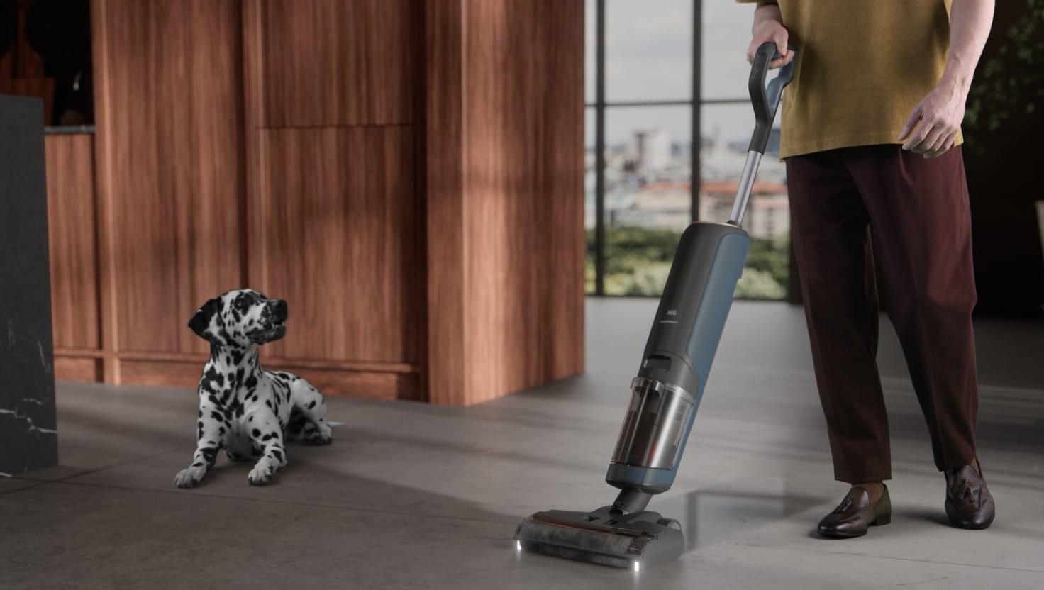 A man cleaning with an AEG stick vacuum cleaner while being watched by a dog lying on the floor.